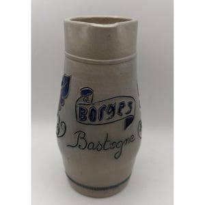 Pode incluir: Um jarro cilíndrico de grés com um acabamento mate em cinzento claro. A parte frontal apresenta uma faixa azul em relevo com o texto "BORGES" e a palavra "Bastogne" escrita em letra cursiva azul por baixo, juntamente com volutas decorativas azuis.
