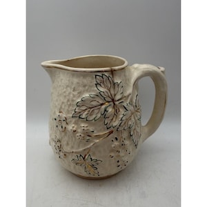 May include: A cream-coloured ceramic pitcher with a textured surface featuring embossed grape leaves and clusters of grapes. The design is accented with hand-painted green outlines and gold metallic detailing along the leaves and fruit.