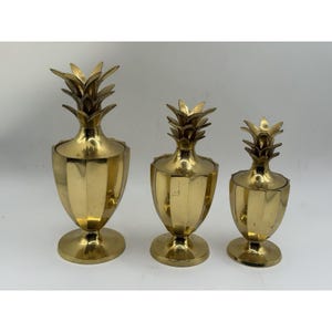 May include: A set of three polished brass containers, each shaped like a pineapple with a removable lid. The containers feature a vertical ribbed body, a circular base, and a textured leaf top, arranged in descending order of height.