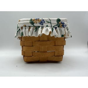 Peut inclure: Un petit panier carré en osier avec une doublure en tissu floral et une bordure à volants. Le panier est en bois brun clair et possède un couvercle en tissu blanc avec des fleurs bleues et jaunes et des feuilles vertes.