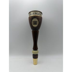 Pode incluir: Uma torneira de cerveja de madeira castanho-escura com uma faixa de metal dourado no topo com o texto ERLANGER impresso a preto. A torneira inclui um emblema oval central com um brasão e um pequeno anel de cor creme sobre a base dourada.