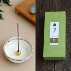 May include: A ceramic incense holder with a gold-coloured ball at the base, holding a dark incense stick. Beside it, a green box with Japanese text and a logo. The box is labelled "Asayu traditional incense Hinoki Made in Japan."