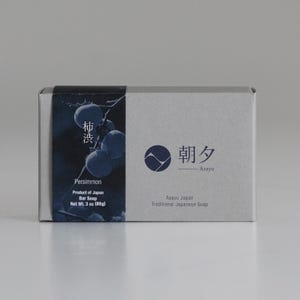 May include: A rectangular box of Asayu Japan Persimmon Bar Soap. The box is light grey with a dark blue panel featuring a persimmon branch illustration and Japanese text. The box is labeled "Net Wt. 85g".