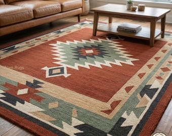 May include: A rectangular area rug with a geometric design. The rug features a central diamond pattern in white, green, and navy blue on a rust-colored background. The border includes tan, green, and black geometric shapes. The rug is placed on a hardwood floor.
