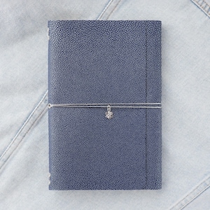 May include: A navy blue leather journal with a textured surface. A silver elastic band wraps around the journal, adorned with a small, decorative charm. The journal is resting on a light blue denim surface.