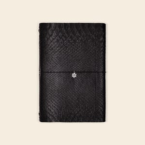 May include: Black leather journal with a textured snakeskin pattern. A black elastic band wraps around the journal, holding it closed. A small, silver-colored flower charm hangs from the elastic band. The journal is rectangular and appears to be closed.