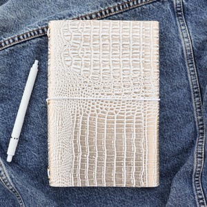 May include: A gold-colored journal with a crocodile skin texture, secured with a white elastic band. A white pen rests beside the journal on a denim jacket.