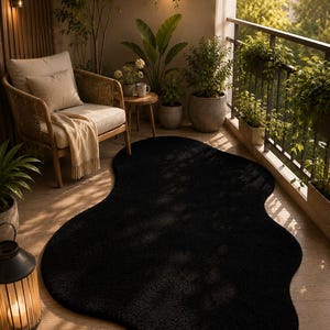 Puede incluir: Una alfombra negra de forma irregular sobre un balcón de madera. La alfombra es el punto focal, con una superficie texturizada. El balcón cuenta con una silla de mimbre, plantas y una barandilla de metal, creando un espacio exterior acogedor.