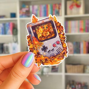 Puede incluir: Una pegatina con un diseño de consola de juegos portátil retro con un personaje Charmander en la pantalla, rodeado de llamas. La pegatina tiene un fondo blanco con botones rojos y negros. El tema son los videojuegos.