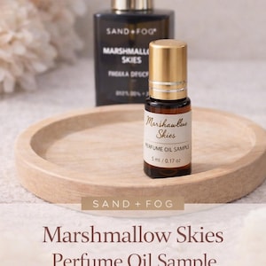 May include: A perfume oil sample bottle with a gold cap sits on a wooden tray. The bottle is labelled "Marshmallow Skies" and contains 5 ml of perfume. A larger black bottle with a gold top and the words "SAND + FOG" is in the background.