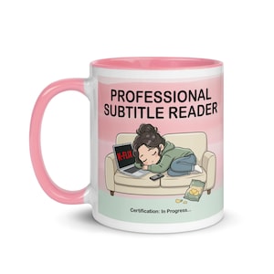 May include: White ceramic mug with a pink interior and handle. The mug features the text "PROFESSIONAL SUBTITLE READER" and an illustration of a person sleeping on a couch with a laptop. The laptop screen displays "K-FLIX".