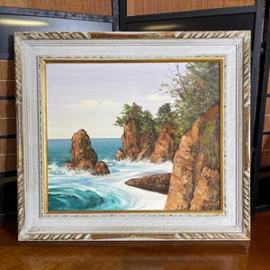 May include: An oil painting of a coastal landscape featuring jagged brown rock formations rising from turquoise ocean waves, with green trees atop the cliffs, all encased in a white wooden frame with a thin gold inner border trim.