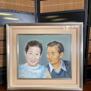 May include: A framed oil painting depicting a portrait of two individuals. The person on the left wears a light blue textured top, while the person on the right wears a dark blue sweater over a white collared shirt. The painting is set in a silver frame.