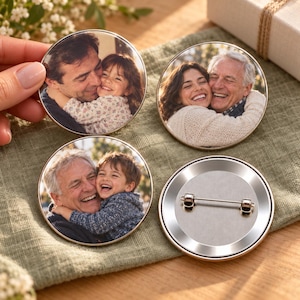 Puede incluir: Tres chapas redondas con retratos fotográficos de familiares abrazándose. Una chapa muestra a un hombre y una niña, otra a una mujer y un hombre mayor, y la tercera a un hombre mayor y un niño pequeño. Una chapa está dada la vuelta para mostrar la parte trasera metálica con un alfiler.