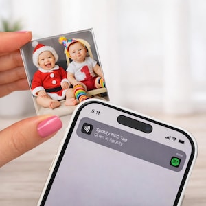 Puede incluir: Un imán fotográfico cuadrado con dos bebés, uno con un traje de Papá Noel y otro con un gorro de fiesta. Un smartphone muestra una notificación: "Spotify NFC Tag Open in Spotify."