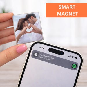 Puede incluir: Imán inteligente cuadrado con una foto de una pareja formando un corazón con sus manos. El imán se sostiene cerca de un teléfono inteligente que muestra una notificación de Spotify NFC Tag. El texto "SMART MAGNET" está en un rectángulo naranja.