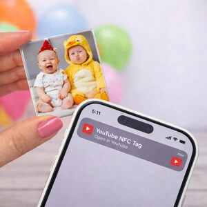 Puede incluir: Impresión fotográfica cuadrada con dos bebés, uno con gorro de fiesta y otro con disfraz de pato amarillo. Un smartphone muestra una notificación de YouTube NFC Tag. Globos de colores en el fondo.