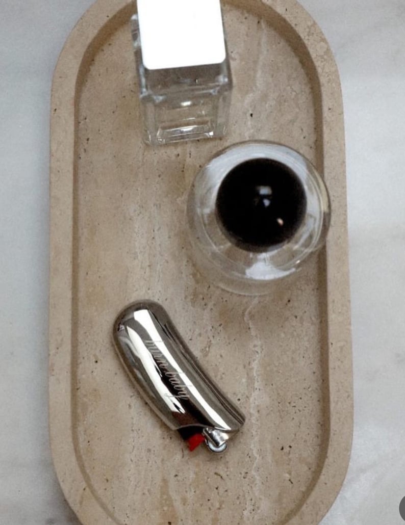 Pu&ograve; includere: Un vassoio ovale beige contiene un accendino argentato con la scritta "mum baby", un contenitore di vetro trasparente e un bicchiere con una sfera nera. L'accendino ha un pulsante rosso.