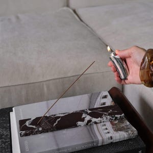 Pu&ograve; includere: Un bastoncino di incenso acceso poggia su un vassoio di marmo posto sopra dei libri. Un accendino d'argento con un pulsante rosso &egrave; tenuto vicino. I libri includono titoli come "Constellation" e "To Breathe". La scena &egrave; ambientata su un tavolo di legno scuro.
