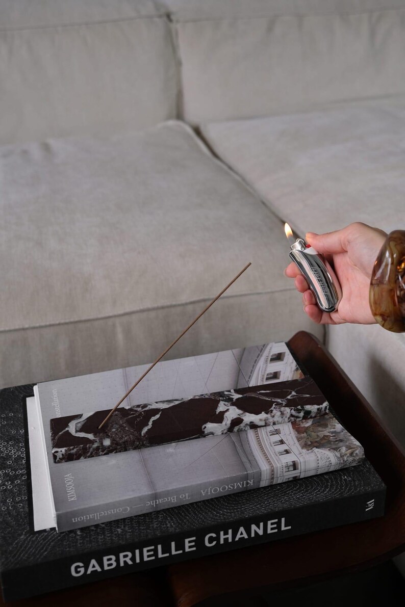 Pu&ograve; includere: Un bastoncino di incenso acceso poggia su un vassoio di marmo posto sopra dei libri. Un accendino d'argento con un pulsante rosso &egrave; tenuto vicino. I libri includono titoli come "Constellation" e "To Breathe". La scena &egrave; ambientata su un tavolo di legno scuro.