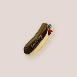 Pu&ograve; includere: Un accendino color oro con la scritta "burn baby" in corsivo. L'accendino ha una forma curva e un meccanismo di accensione rosso e nero. Lo sfondo &egrave; di colore rosa chiaro uniforme.