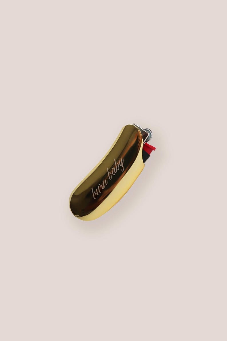 Pu&ograve; includere: Un accendino color oro con la scritta "burn baby" in corsivo. L'accendino ha una forma curva e un meccanismo di accensione rosso e nero. Lo sfondo &egrave; di colore rosa chiaro uniforme.