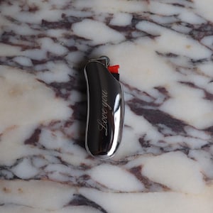 Pu&ograve; includere: Un accendino cromato con la scritta "Love you" incisa sul lato. L'accendino ha un pulsante di accensione rosso ed &egrave; posto su uno sfondo di marmo.