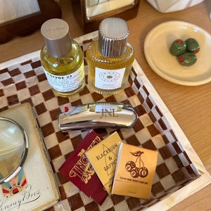 以下が含まれることがあります： 金色の液体が入った2つのガラス製香水瓶（Le Labo Poivre 23とJorum Studio Sunset Vanillaのラベル付き）。JNLの刻印がある銀色のライター、3つの小さなマッチ箱、虫眼鏡が、茶色と白の市松模様のトレイの上に置かれています。