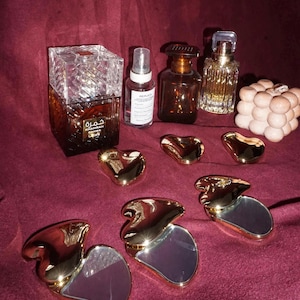Peut inclure: Une collection de quatre flacons de parfum, dont un flacon carré étiqueté Khamrah Qahwa, un flacon pulvérisateur transparent, un flacon ambré foncé et un flacon doré, avec une bougie bulle beige et six miroirs dorés en forme de cœur.