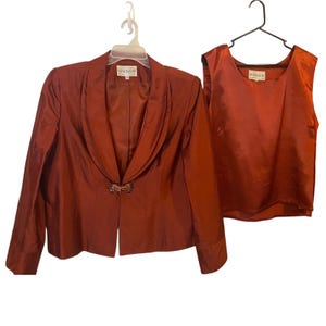 May include: A rust-colored two-piece set consisting of a long-sleeved jacket and a sleeveless shell top. The jacket features a shawl collar and a single decorative clasp closure. Both garments have a satin-like finish and Lily & Taylor labels.