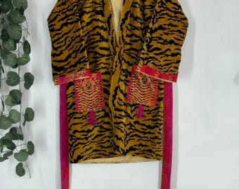 May include: A mustard yellow robe with black tiger stripes, a striped interior, and bright pink trim. The robe has two pockets with orange and black tiger print and pink tassels. The robe is hanging on a wooden hanger.