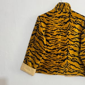 May include: A gold and black tiger-striped jacket with a high collar and long sleeves. The cuffs are lined with a cream and yellow striped fabric. The jacket is hanging on a metal hanger against a white wall.