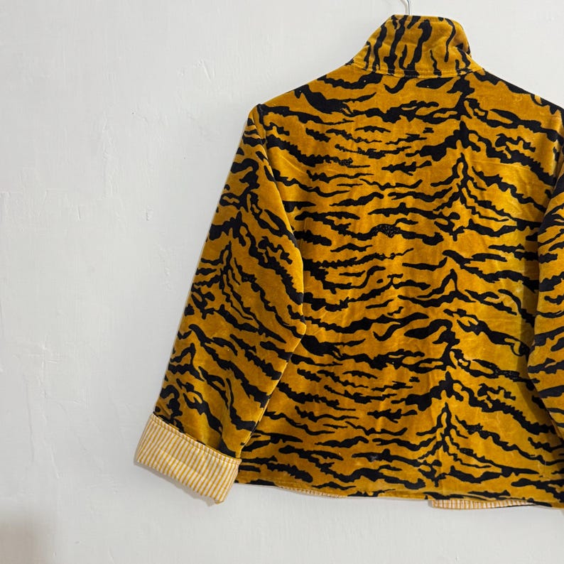 May include: A gold and black tiger-striped jacket with a high collar and long sleeves. The cuffs are lined with a cream and yellow striped fabric. The jacket is hanging on a metal hanger against a white wall.