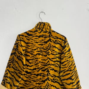 May include: A gold and black tiger-striped jacket hanging on a metal hanger. The jacket has a high collar and long sleeves. The fabric has a soft, velvety texture. The cuffs have a striped pattern.