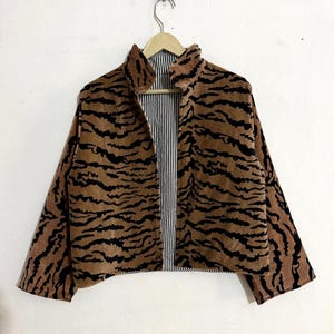 Könnte beinhalten: Eine kurz geschnittene Jacke mit einem braun-schwarzen Tigerstreifenmuster auf der Außenseite. Das Kleidungsstück hat einen Stehkragen, lange Ärmel und eine offene Vorderseite, die ein schwarz-weißes vertikales Nadelstreifenfutter zeigt.