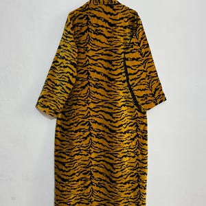 May include: A long, open-front robe with a striking tiger stripe pattern in shades of gold and black. The robe is hanging on a metal hanger against a plain white wall. The garment has long sleeves and a knee-length cut.