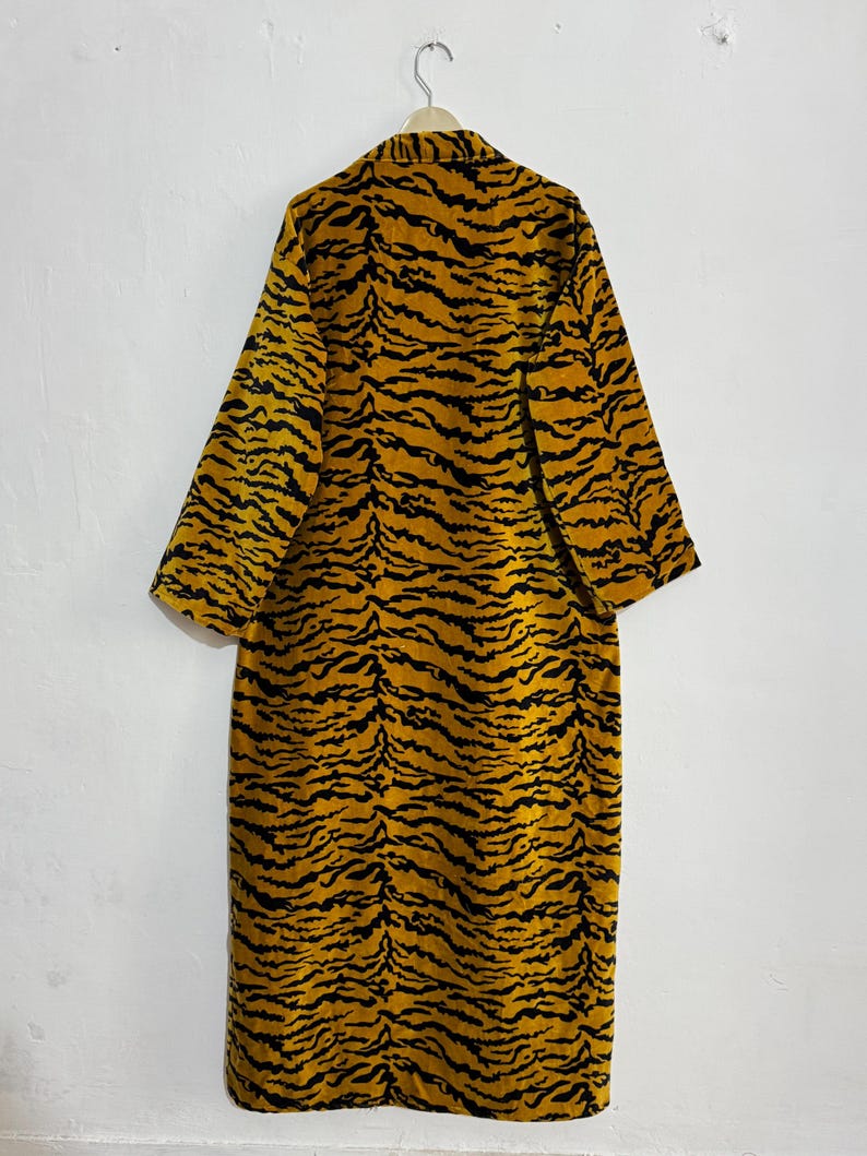 May include: A long, open-front robe with a striking tiger stripe pattern in shades of gold and black. The robe is hanging on a metal hanger against a plain white wall. The garment has long sleeves and a knee-length cut.