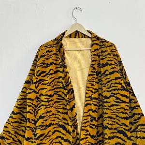 May include: A yellow and black tiger-striped robe hanging on a wooden hanger. The robe has a soft, velvety texture and a contrasting yellow and white striped lining. The robe's design mimics a tiger's pattern.