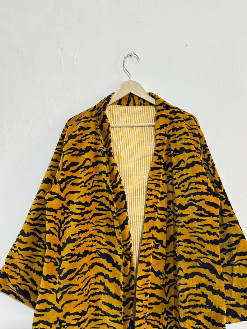 May include: A yellow and black tiger-striped robe hanging on a wooden hanger. The robe has a soft, velvety texture and a contrasting yellow and white striped lining. The robe's design mimics a tiger's pattern.