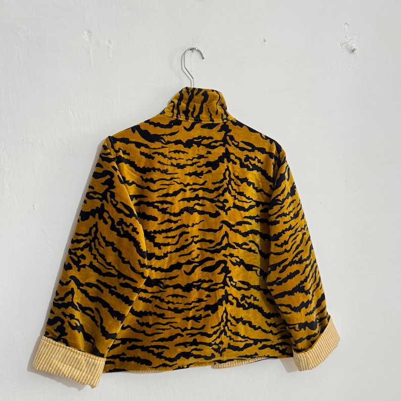 May include: A mustard yellow jacket with a black tiger stripe pattern. The jacket has a high collar and folded cuffs with a cream and yellow striped lining. The jacket is hanging on a silver hanger against a white wall.