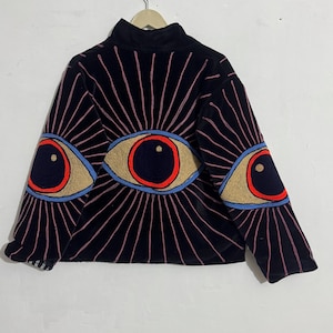 May include: Black velvet sweatshirt with embroidered eye designs. The sweatshirt features three large eye motifs on the sleeves and chest, with red, tan, blue, and pink accents. The design includes radiating lines emanating from the eyes.