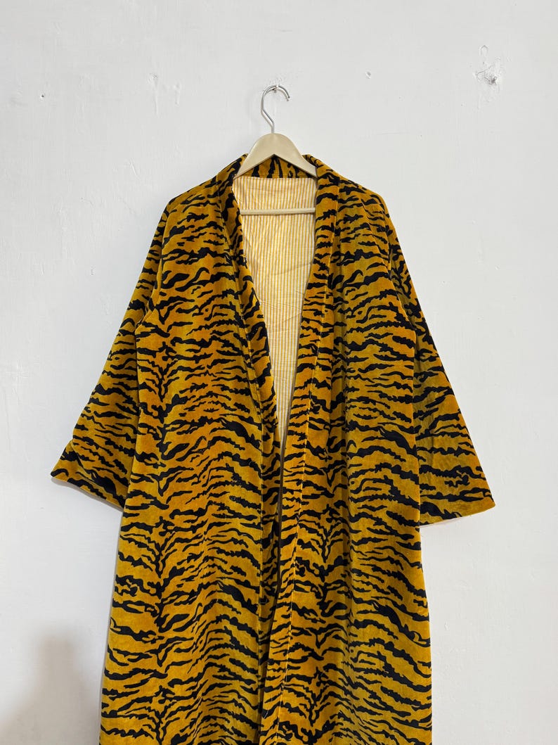 May include: A long, open-front robe with a tiger stripe pattern in black and yellow. The robe is hanging on a wooden hanger against a white background. The interior lining is yellow and white striped.