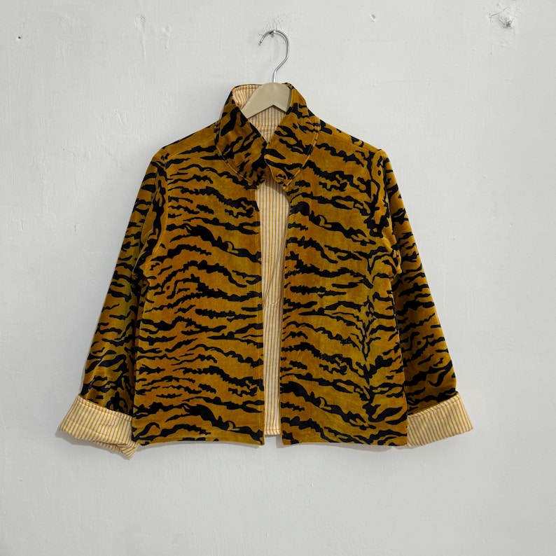 May include: A mustard yellow jacket with a black tiger stripe pattern. The jacket has a collared neckline and long sleeves with striped cuffs. The interior lining is a light yellow and white striped fabric.