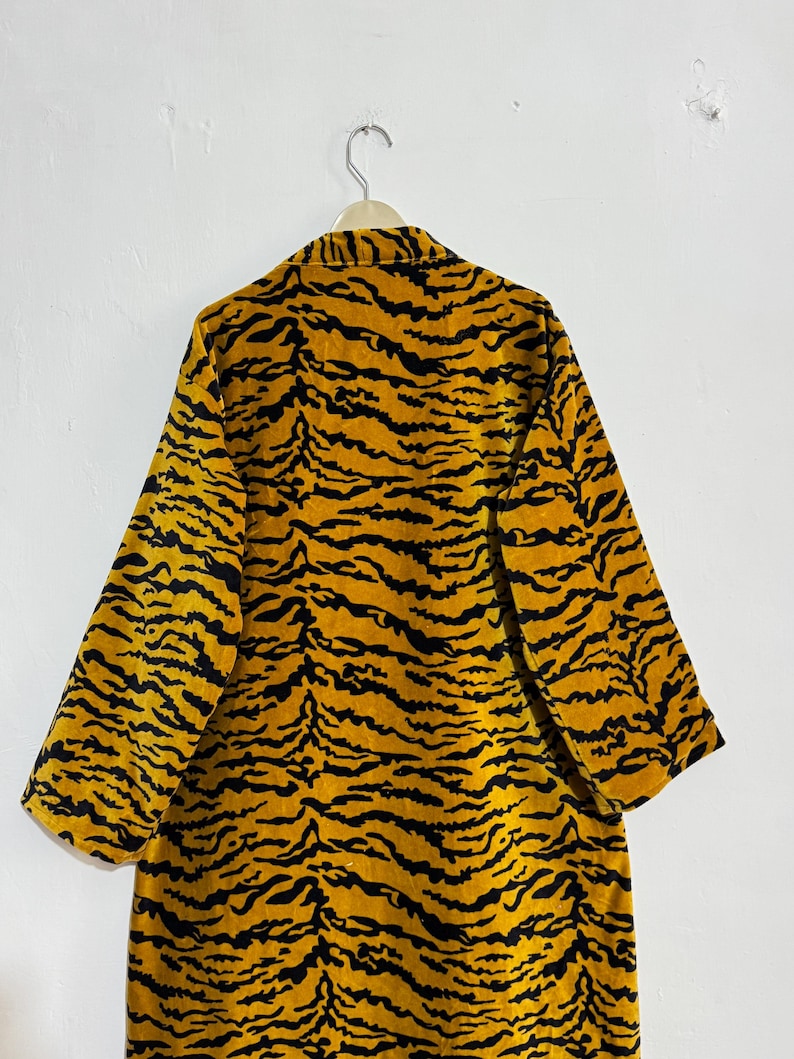 May include: A long, open-front robe with a tiger stripe pattern in shades of yellow and black. The robe is hanging on a hanger against a white wall. The sleeves are wide and three-quarter length.