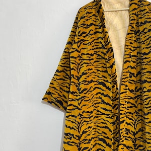 May include: A long, open-front robe with a vibrant tiger stripe pattern in black and yellow. The robe is hanging on a wooden hanger against a white wall. The interior lining is a light yellow with vertical stripes.