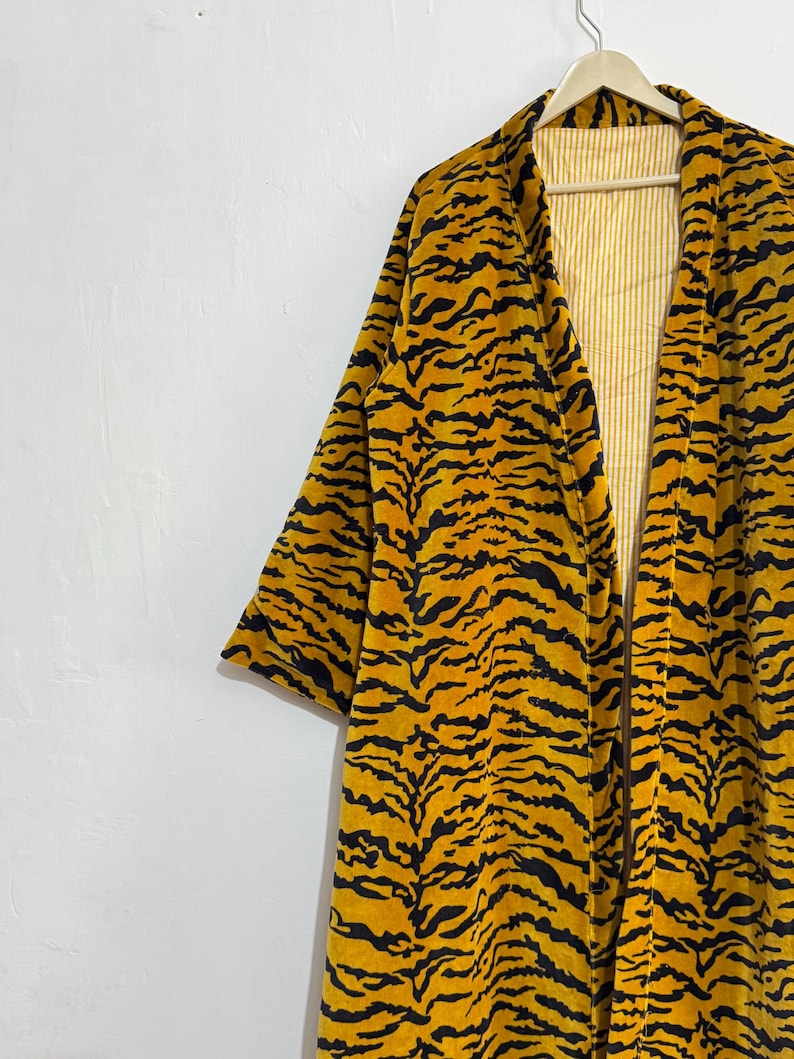 May include: A long, open-front robe with a vibrant tiger stripe pattern in black and yellow. The robe is hanging on a wooden hanger against a white wall. The interior lining is a light yellow with vertical stripes.