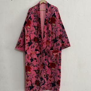 Peut inclure: Robe rose à imprimé floral avec un motif rouge et bleu marine. La robe a des manches longues, deux poches avant et un intérieur rayé. Elle est suspendue à un cintre en bois contre un mur blanc.