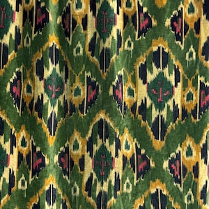 Op de afbeelding: Close-up van een groene fluwelen stof met een herhalend geometrisch patroon. Het ontwerp bevat ruitvormen in tinten groen, goud, zwart en roze. De stof heeft een zachte, getextureerde uitstraling, wat suggereert dat deze gebruikt kan worden voor bekleding of decoratieve doeleinden.