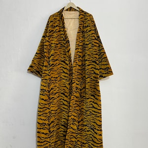 May include: A long, open-front robe with a tiger stripe pattern in shades of yellow and black. The robe is hanging on a wooden hanger against a white wall. The interior lining is a light yellow with vertical stripes.