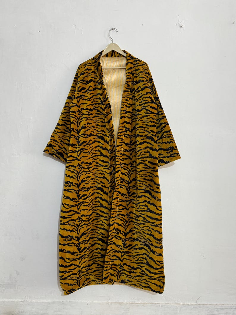 May include: A long, open-front robe with a tiger stripe pattern in shades of yellow and black. The robe is hanging on a wooden hanger against a white wall. The interior lining is a light yellow with vertical stripes.
